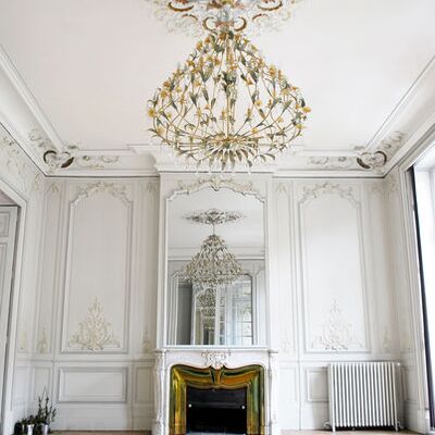 Grand lustre floral, cheminée dorée et moulures sur les murs et plafond. Parquet en bois clair.