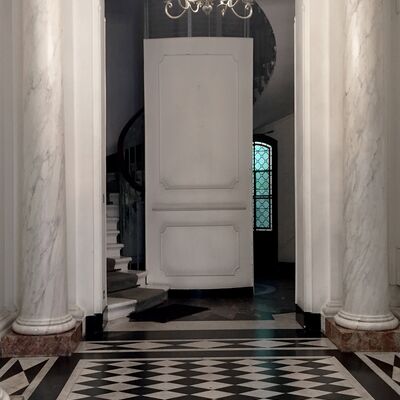 Sol en damier noir et blanc. Colonnes en marbre de chaque côté. Lustre au plafond et escalier circulaire blanc en fond.